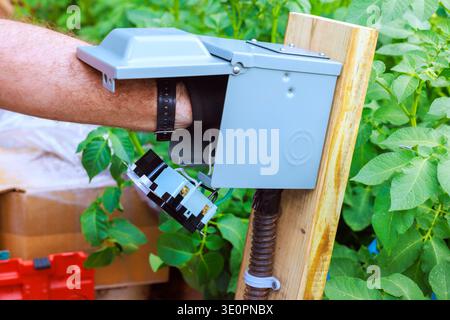 Person uses their hand to install electrical parts in gray box on post surrounded by greenery Stock Photo