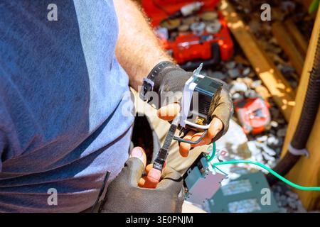 Worker is holding tool, connecting wires while sitting in workshop full of tools materials scattered around Stock Photo