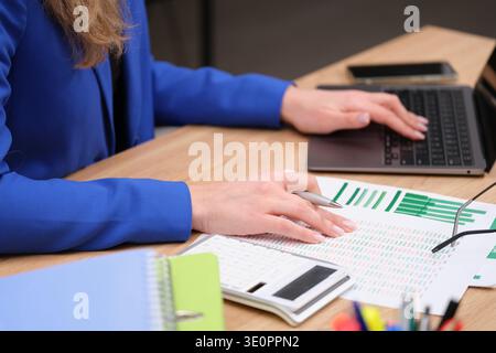 Professional woman analyzing data and working on a laptop while surrounded by charts and office supplies during a productive workday Stock Photo