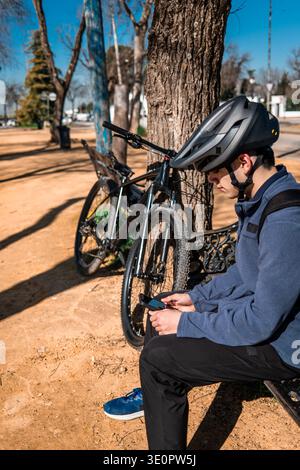 Young man wearing helmet sitting on a bench in a park, using his cell phone with his mountain bike nearby Stock Photo