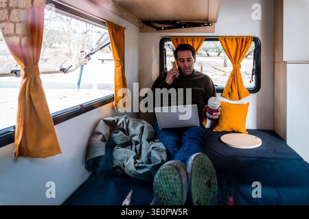Man working with laptop and talking on mobile phone, living the digital nomad lifestyle in a camper van Stock Photo