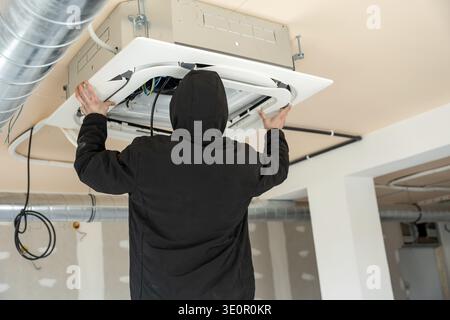 Hvac professional installs a modern air conditioning ceiling cassette unit on a construction site, connecting wiring and adjusting components for vent Stock Photo