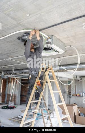 Skilled technician standing on a ladder and connecting pipes and wiring to a new air conditioning unit, working on the ceiling of a building construct Stock Photo
