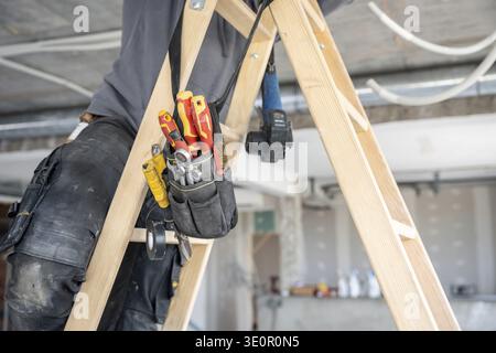 Construction worker wearing a tool belt filled with various hand tools, standing on a ladder and performing tasks during the renovation of a building Stock Photo