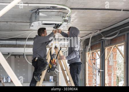 Hvac technicians working on a ladder, connecting wires to an air conditioning system mounted on the concrete ceiling during construction of a modern r Stock Photo