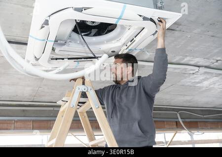 Professional hvac technician installing a ceiling mounted air conditioning unit on a ladder, working to integrate the system into a building under con Stock Photo