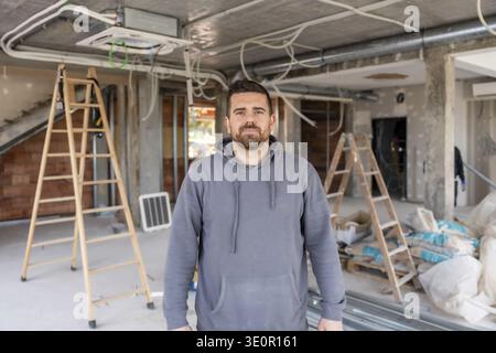 Male professional installer or construction worker standing in an unfinished building, surrounded by ladders, air conditioning units, and renovation m Stock Photo