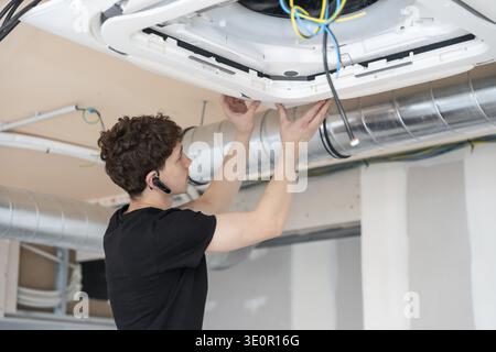 Young male technician installing a modern cassette air conditioning unit into a ceiling during commercial or residential construction, connecting wire Stock Photo