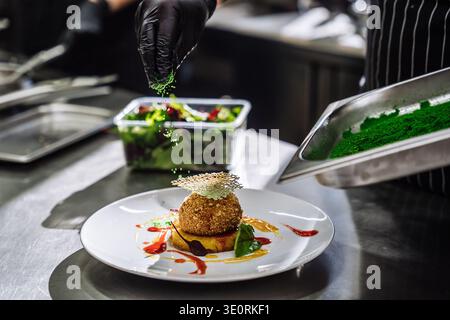 A close up shot of a chef's hand in a black nitrile glove sprinkling fine green dust over a breaded dessert ball. The dessert is beautifully plated on Stock Photo