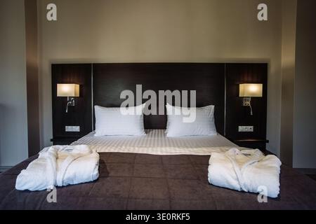 A wide shot of a neatly made large bed with a dark wood headboard and symmetrical night lamps. Two white folded bathrobes lie on the brown bedspread i Stock Photo