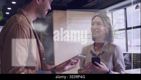 Holding phone, woman in blouse checking data on glass wall while coworker holding tablet in office Stock Photo
