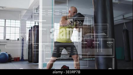 Man wearing yellow top, black shorts, boxing gloves punching heavy bag in gym, with HUD overlay Stock Photo