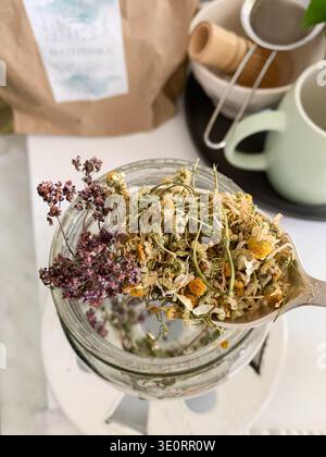 A person holds a spoonful of dried calendula and chamomile flowers, ready to make herbal tea. The kitchen has jars, a bag of herbs, and tea cups on a Stock Photo