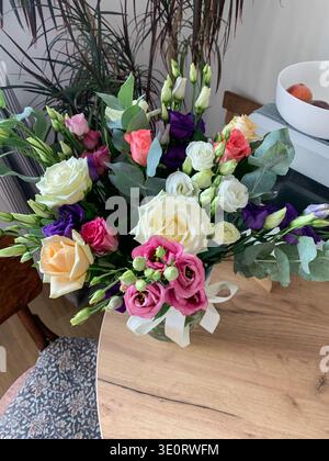 A flower arrangement sits on a wooden table. The bouquet has several types of flowers in different colors. Nearby, a bowl holds fruit and plants adorn Stock Photo