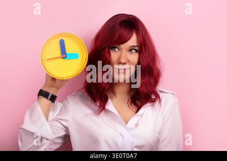 Confident woman with red hair holding a yellow clock against a pink background, wearing a white shirt and black watch, symbolizes time management and Stock Photo