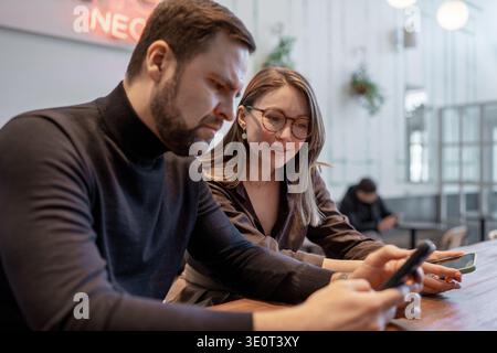 Young colleagues laughing and sharing smartphones at high table in bright open cafe. Laughing, smartphones, high table, cafe, teamwork. Stock Photo