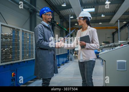 Female factory manager and industrial engineer shake hands while discussing machinery and operations in a manufacturing plant. Stock Photo