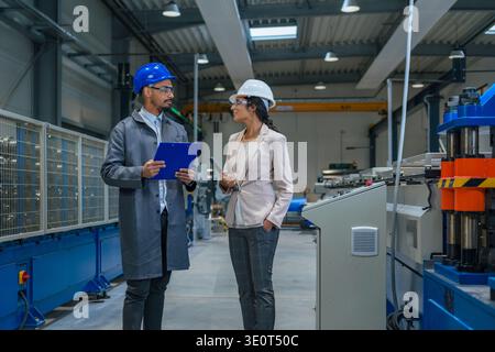 Female factory manager and industrial engineer discuss machinery and operations in a modern manufacturing plant during working hours. Stock Photo