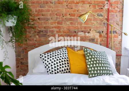Cozy modern bedroom interior design with white bed, decorative pillows, golden lamp and brick wall. Stylish loft style home design with plants Stock Photo