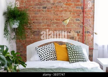 Cozy modern bedroom interior design with white bed, decorative pillows, golden lamp and brick wall. Stylish loft style home design with plants Stock Photo
