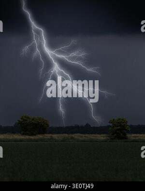 Lightning strike over farm field with trees under dark rain clouds Stock Photo