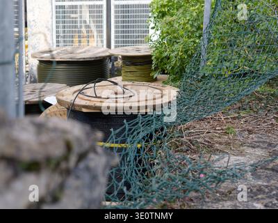 Large spools of black and yellow cable are stored outdoors behind a green chain-link fence, suggesting an infrastructure project or utility work. Stock Photo