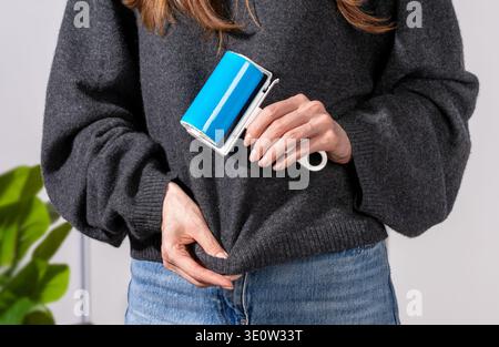 Woman cleaning dark sweater with reusable adhesive sticky roller removing pet hair. Concept of minimizing lint on black clothes, tidy appearance and e Stock Photo