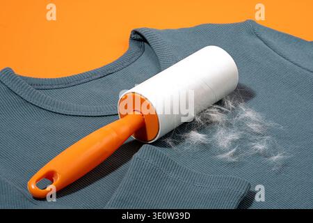 Adhesive lint roller removing pet hair from teal blue sweater on bright orange background. Concept of sticky tape solution for shedding control, cloth Stock Photo