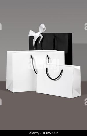 Three paper shopping bags, white and black with handles on grey background. Concept of premium gifting, stylish packaging, holiday shopping display an Stock Photo