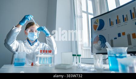 A young female scientist performs chemical experiments in a bright laboratory, using pipettes and test tubes. Data analysis and scientific research are displayed on a computer monitor nearby Stock Photo