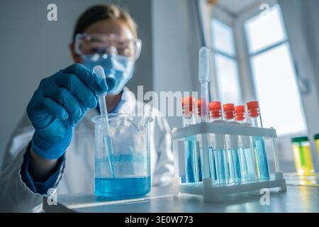 A researcher wearing gloves and safety gear uses a pipette to transfer blue solution into a beaker in a modern lab. Sunlight streams through the window, illuminating scientific equipment and glassware Stock Photo