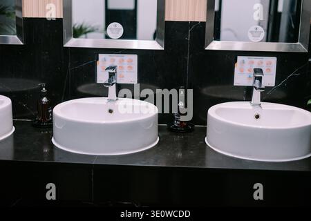 A clean, modern public washroom features multiple white ceramic sinks, chrome faucets, and soap dispensers. Clear signs with handwashing instructions Stock Photo