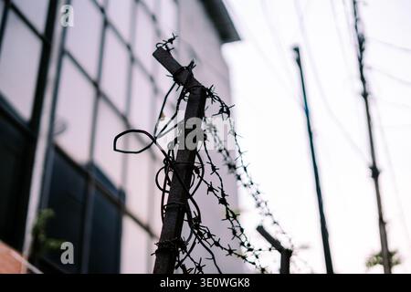 Close-up of rusty barbed wire wrapped around a metal pole, standing as a security fence with a blurred urban building in the desaturated background, s Stock Photo