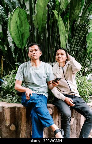 Two men are sitting casually on an outdoor wall, surrounded by vibrant green tropical plants. One man looks at the camera, while the other checks his Stock Photo