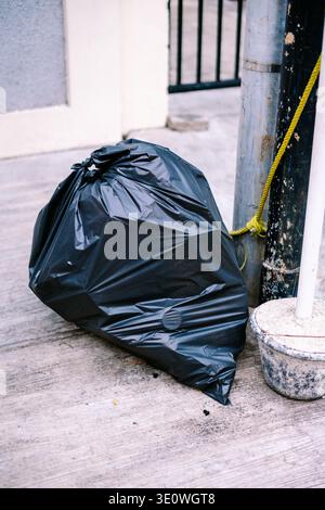 A large, black, tied plastic garbage bag rests on a textured concrete pavement next to weathered utility poles, a yellow rope, and a building wall in Stock Photo
