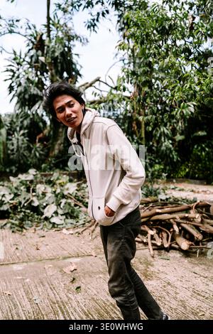 A young man wearing a casual light-colored hoodie stands outdoors with hands in pockets. Surrounded by lush green trees and trimmed branches, he conve Stock Photo