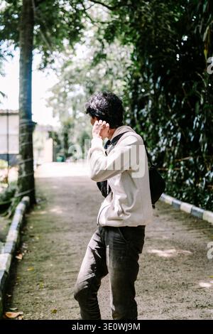 A young man walks casually along a sunny outdoor path, surrounded by lush green foliage. He appears relaxed, enjoying the tranquil environment and bri Stock Photo