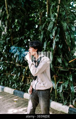 A young Asian man in a casual outfit enjoys a smoke break outdoors. He exhales blue smoke against a backdrop of vibrant green foliage, appearing relax Stock Photo