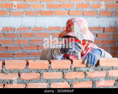 A construction worker wearing a protective sun hat and mask, carefully applying mortar with a trowel while laying red bricks to build a wall. Stock Photo