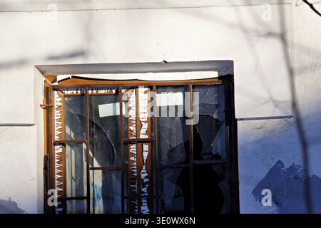 A neglected building facade featuring a damaged window with shattered glass panes contrasting against a bright, textured white wall Stock Photo