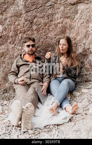 Couple beach picnic man and woman sitting on blanket eating snacks against rock wall with a laptop during vacation Stock Photo