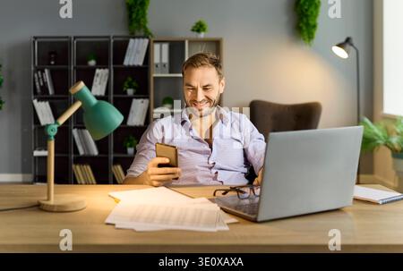 Smiling business man reading message on phone working with laptop in office. Stock Photo