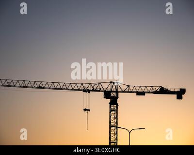 Tower crane silhouette against gradient sunset sky with hanging hook and streetlight in Estonia, March 2026 Stock Photo