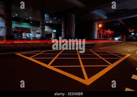Long exposure captures red light trails of vehicles under Taipei MRT pillars at night, with station signs, streetlights, and diamond road markings. Stock Photo