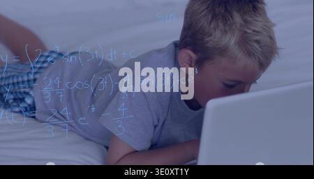 Lying child using laptop in bedroom on bed, wearing gray tee, checkered shorts, blue math overlay Stock Photo