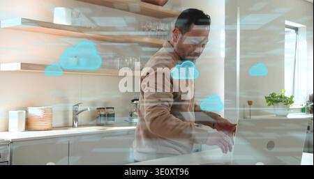 Standing Asian man in beige pullover working on laptop at kitchen counter, showing cloud overlays Stock Photo