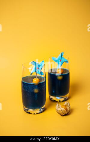 Two glasses of deep blue liquid decorated with pinwheels and physalis sit on yellow background. physalis cocktail garnish, cape gooseberry drinks Stock Photo