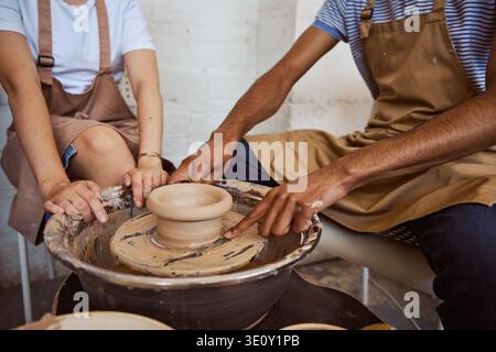 Two focused adults shape wet clay on a pottery wheel in a bright studio. Their collaboration highlights creativity and the joy of crafting ceramic bow Stock Photo
