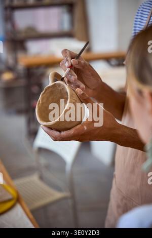 Young man painting clay mug in pottery studio with woman observing Stock Photo