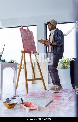 Male artist standing in bright art studio working at wooden easel with paint palette, copy space Stock Photo